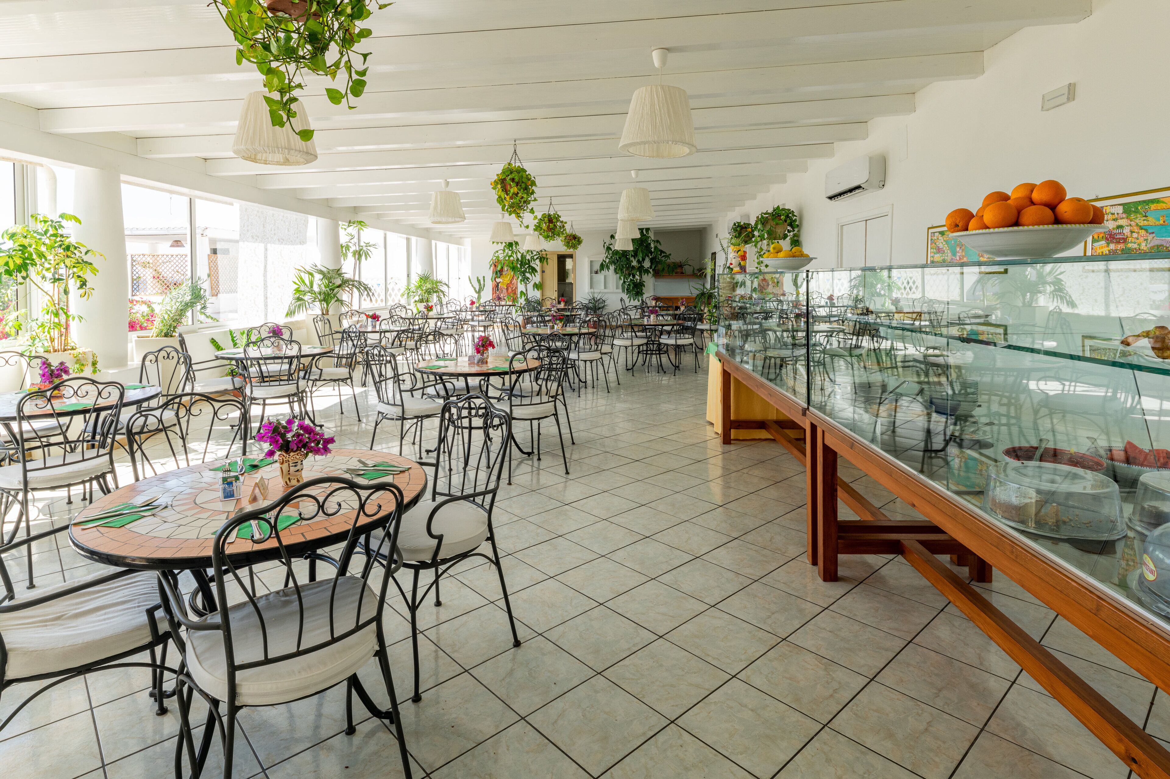 En rummelig café med flere borde og stole, potteplanter og en glasmontre med mad.