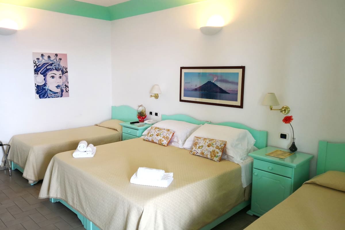 Habitación de hotel con dos camas individuales, una cama grande, un cuadro de un volcán, una fotografía enmarcada y una mesita con una lámpara.