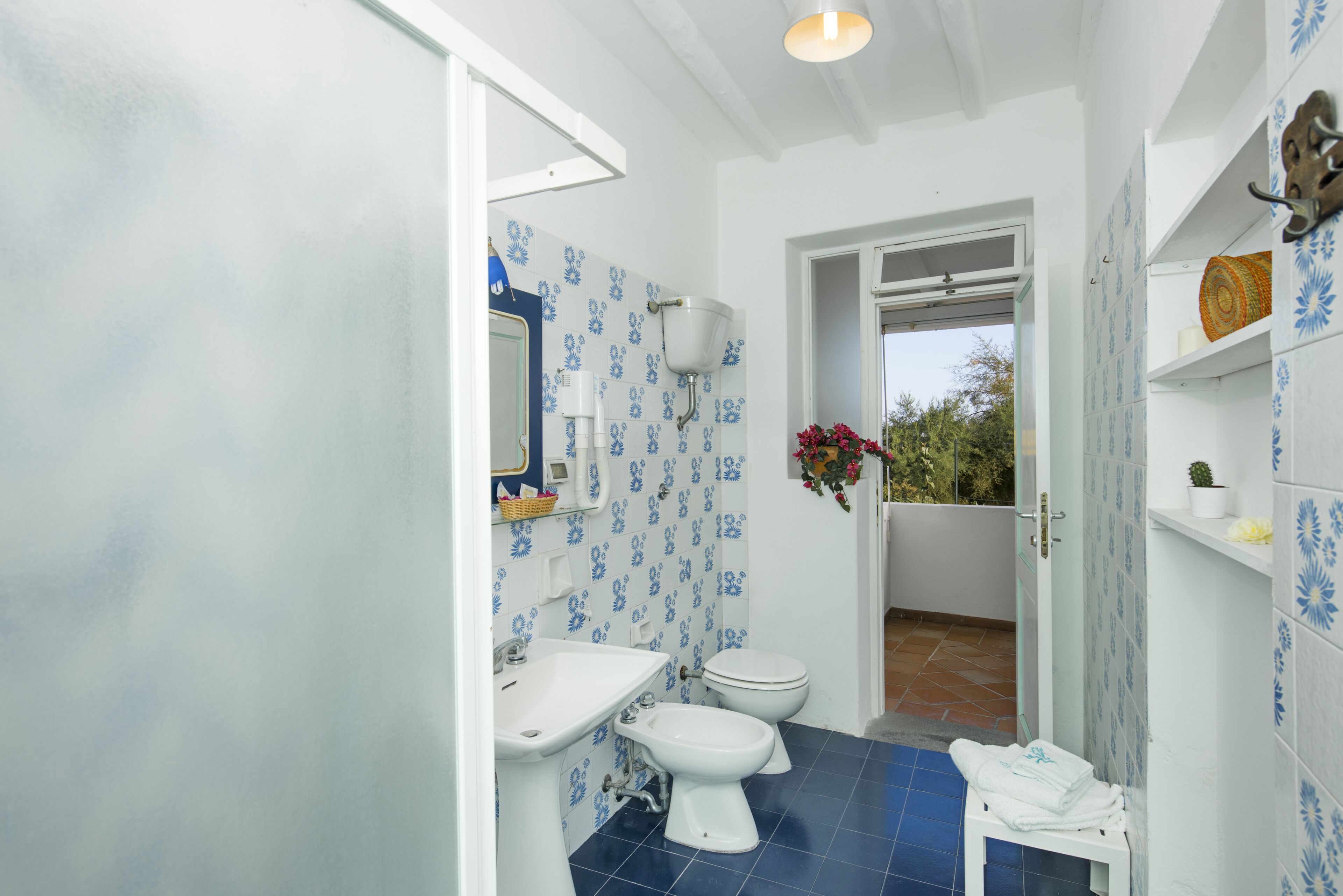 Une salle de bain équipée d’un lavabo, de toilettes et d’une douche. Une fenêtre offre une vue sur la verdure environnante.