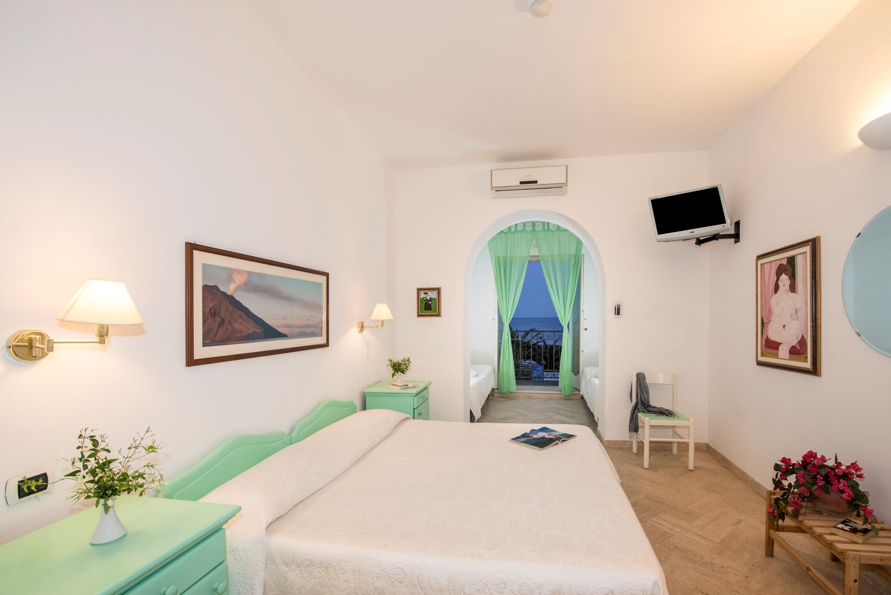 A bedroom with a bed, a nightstand with a lamp, a TV mounted on the wall, and a window with a view of the outside.