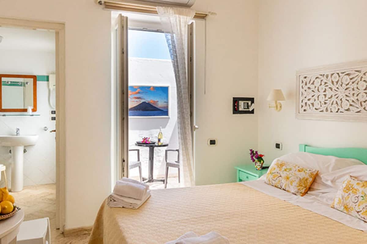 Habitación de hotel con cama, mesita de noche con un cesto de limones, baño con lavabo y espejo, y balcón con vistas al mar.