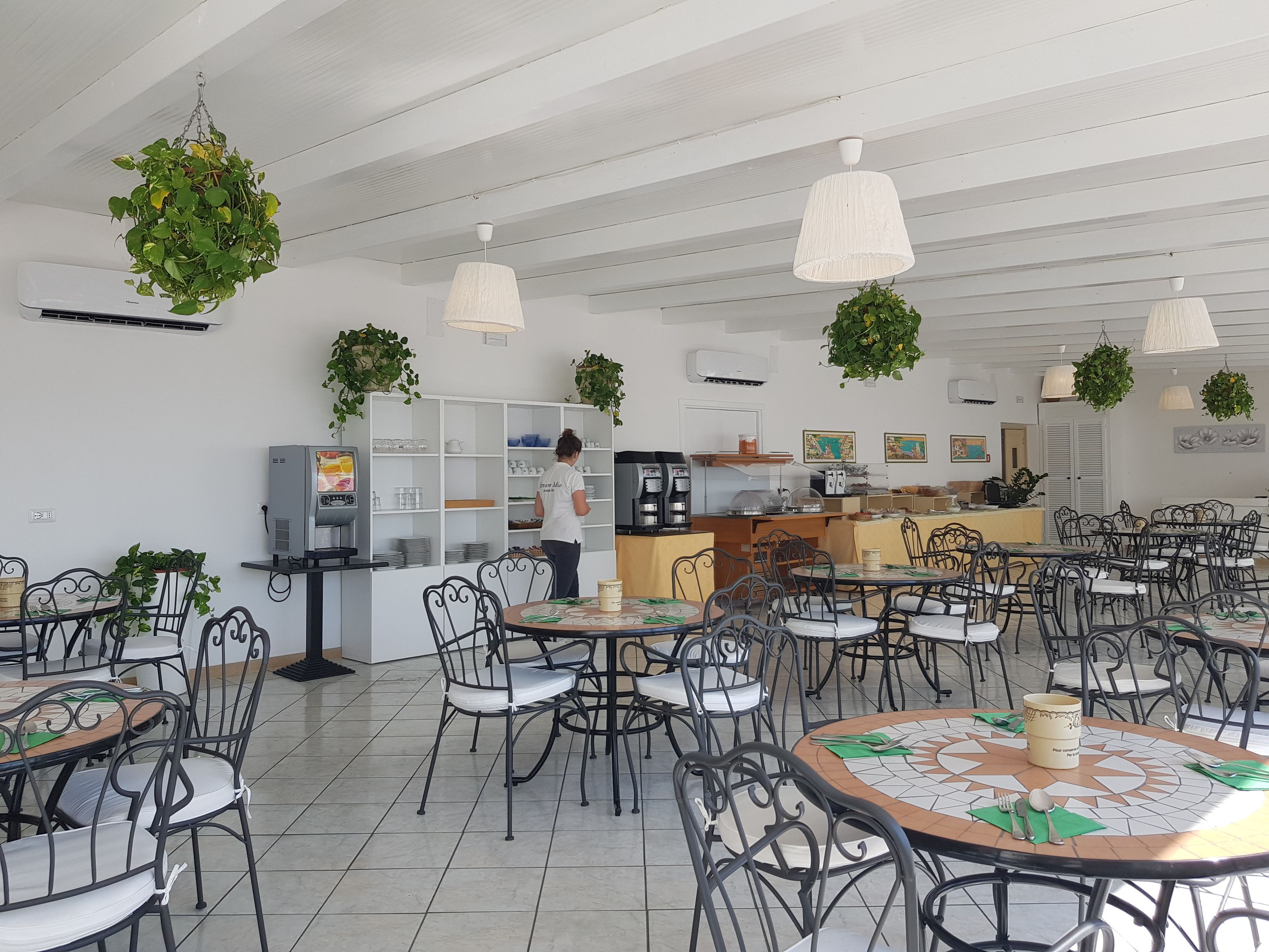 Un restaurant avec des tables rondes et des chaises en métal noir, un coin bar et des plantes suspendues.