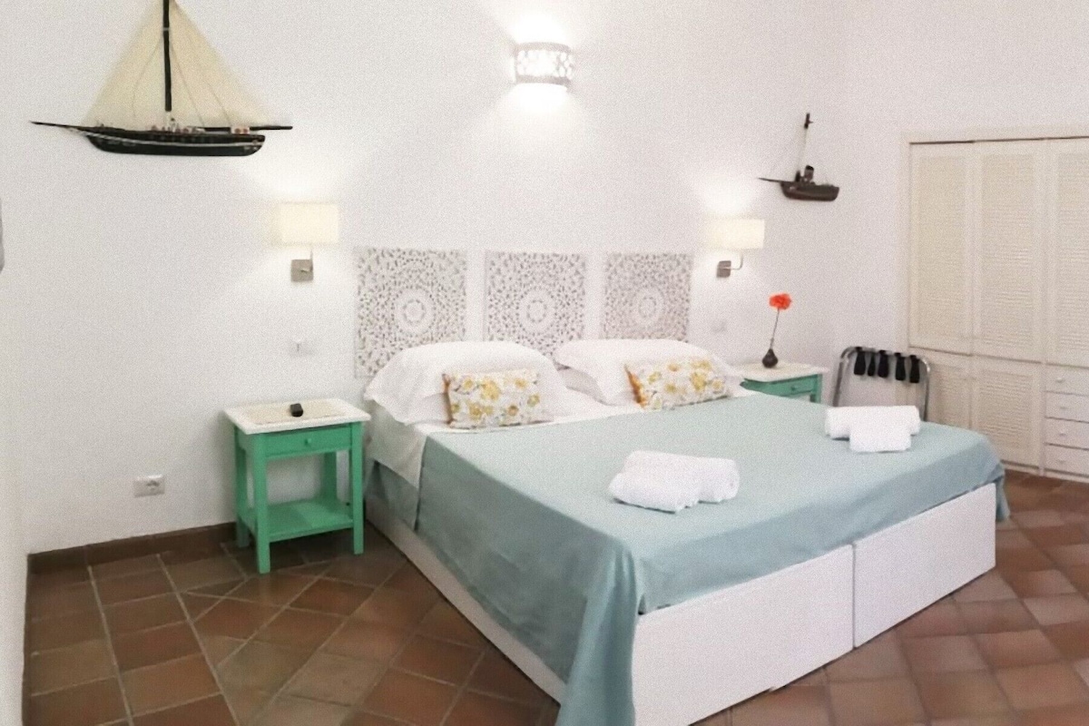 Un dormitorio con cama, mesitas de noche, lámpara y ventana con cortinas.
