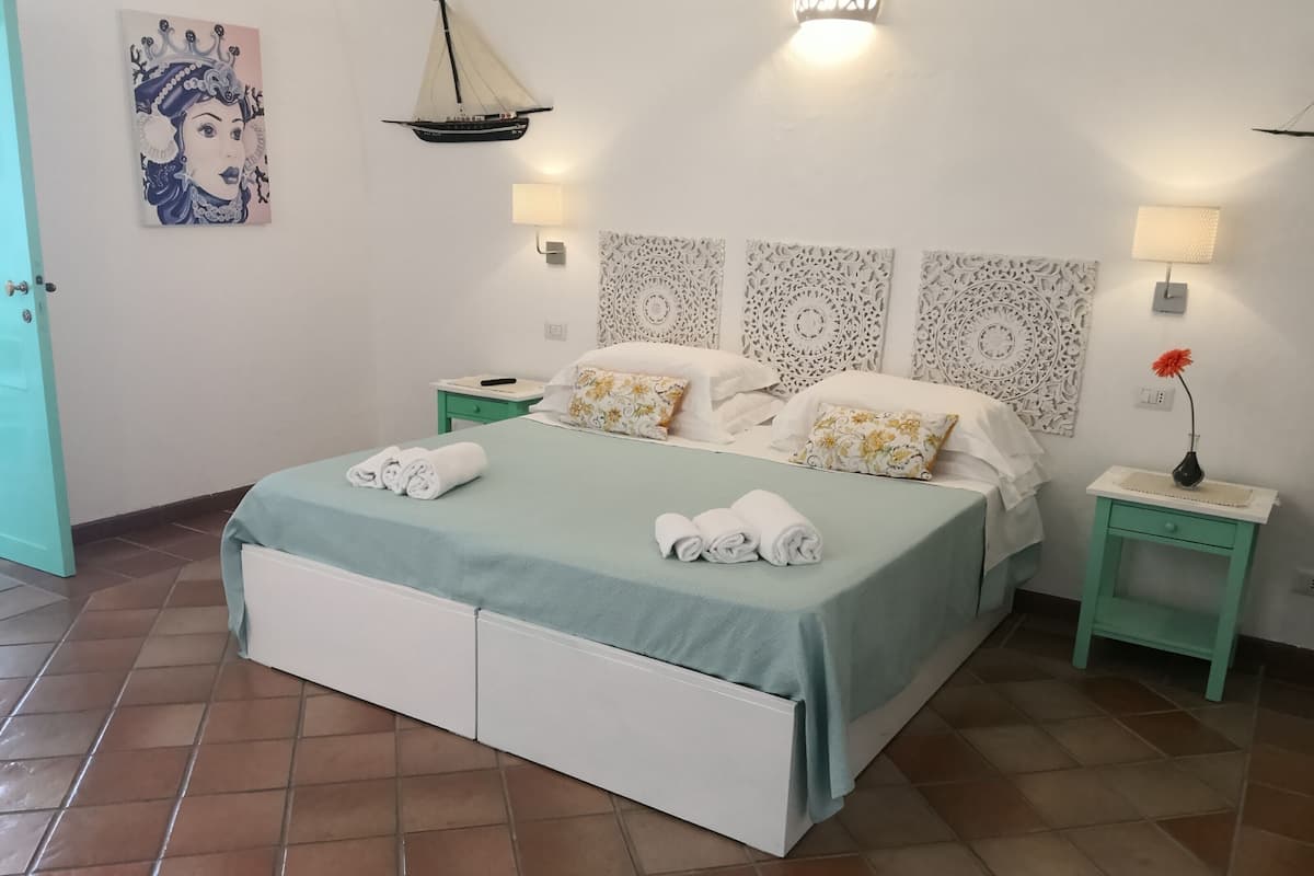Una cama bien hecha con una colcha azul claro y almohadas blancas, una mesita de noche con una lámpara, y un adorno decorativo sobre la cama.