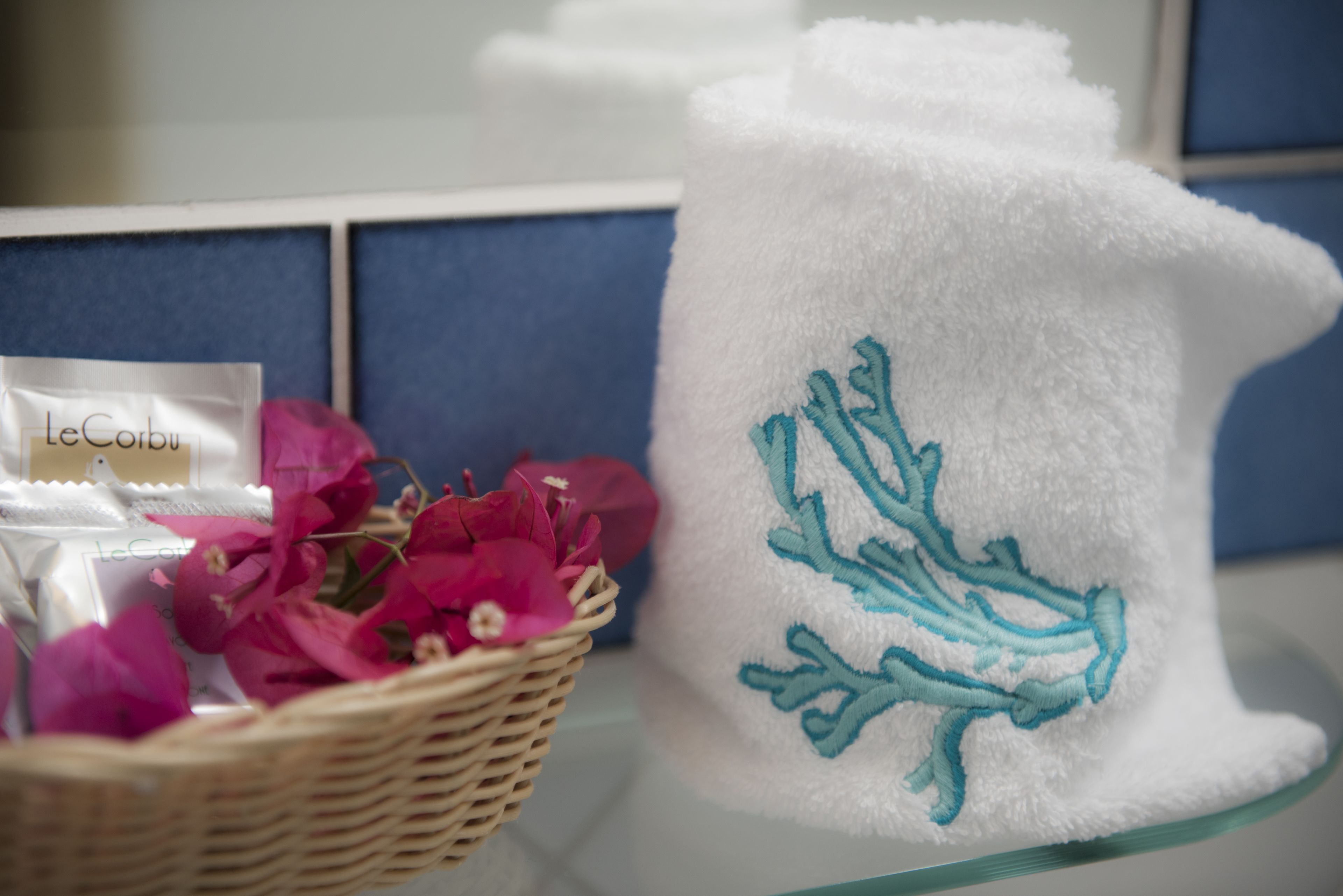 Un asciugamano bianco con ricami blu, un cesto di vimini con fiori rosa e articoli a marchio LeCorbusier.