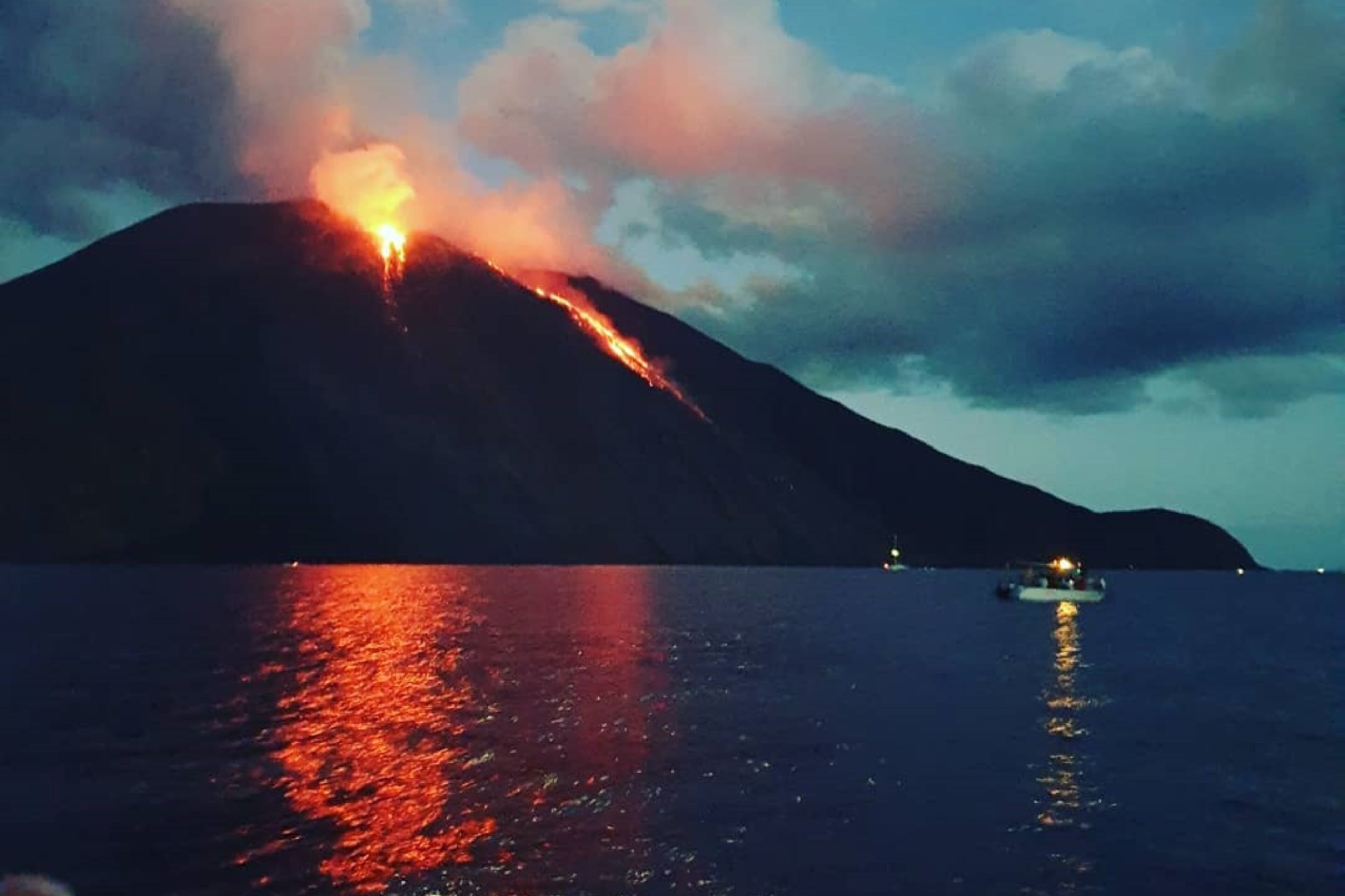 A volcano erupting at night with lava flowing down its side, reflecting on the water.