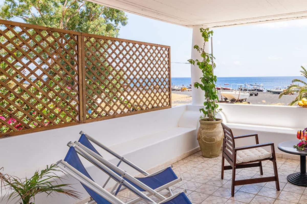 Un balcón con vista a la playa, dos sillas, una mesa y plantas en macetas.