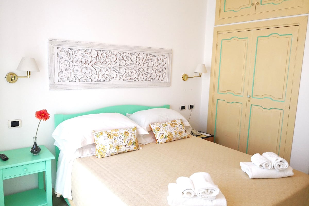 Una cama bien hecha con almohadas florales, un florero con una flor roja y toallas enrolladas sobre la cama.