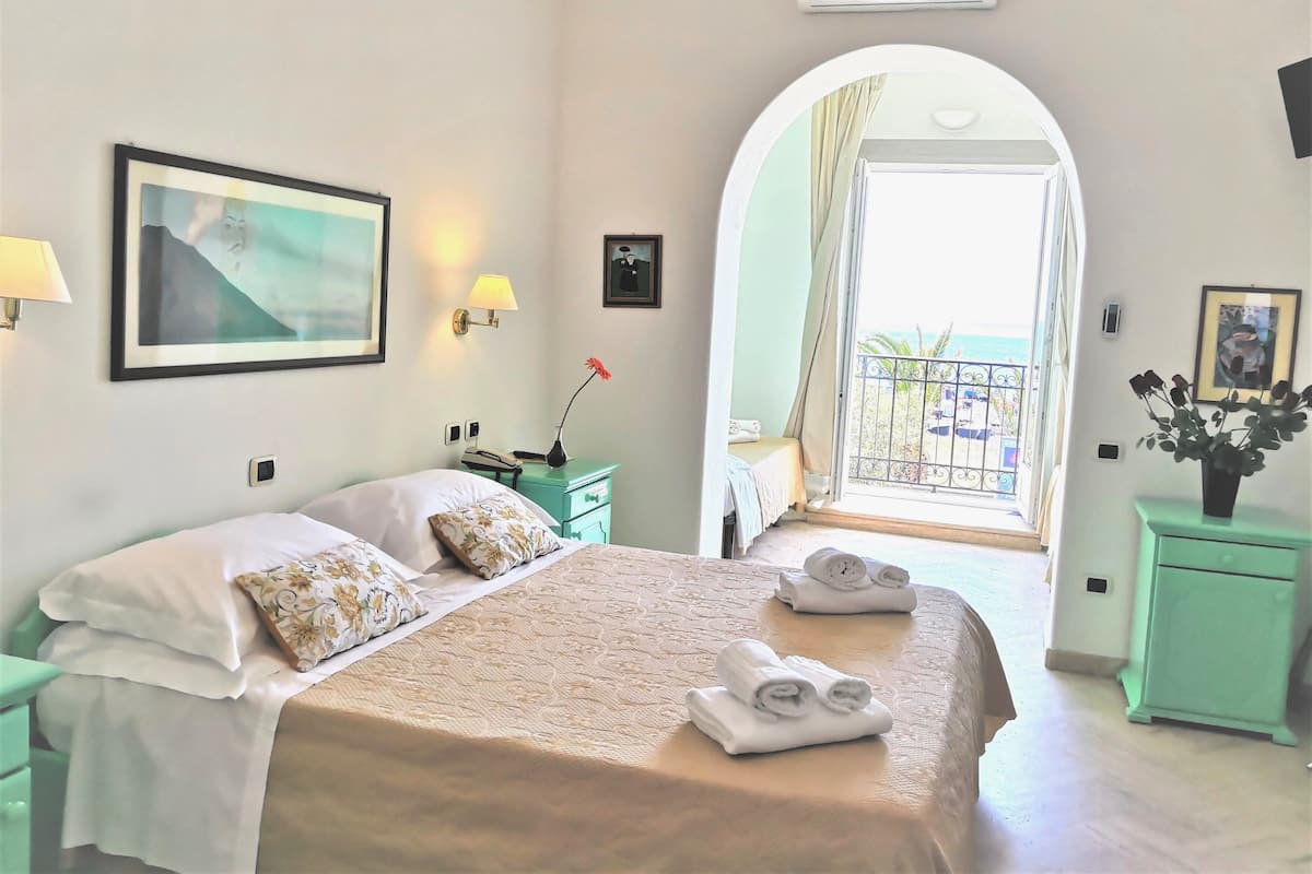 Habitación de hotel con cama, mesita de noche, lámpara, cuadro enmarcado, planta en maceta y balcón con vista a la playa.