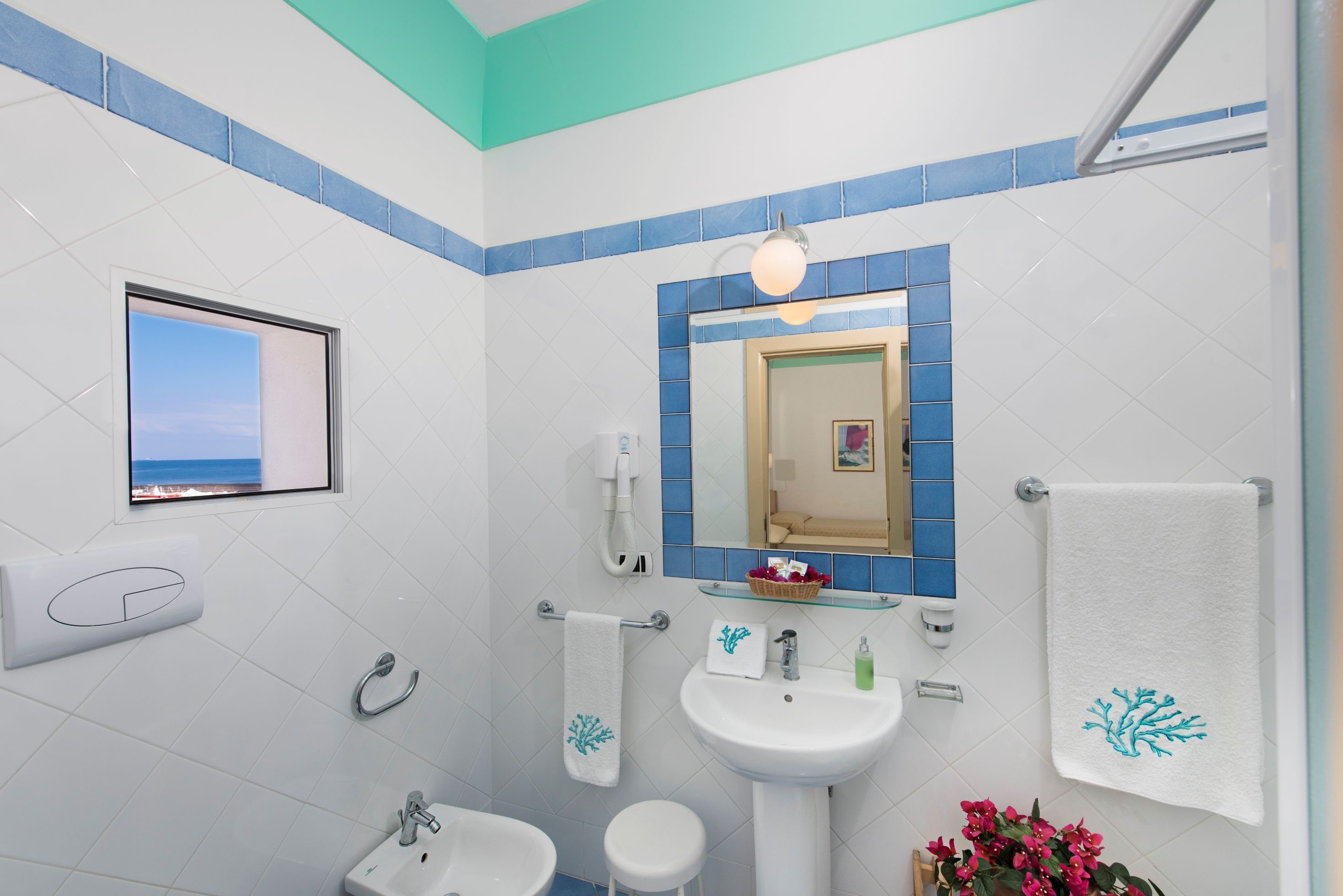Ein Badezimmer mit weißem Waschbecken, einem Spiegel mit blauem Rahmen, einem Handtuchhalter und einem Fenster mit Meerblick.