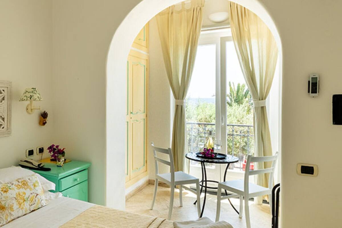 Un dormitorio con cama, una mesita de noche verde, una mesa con sillas y vistas a través de un arco.