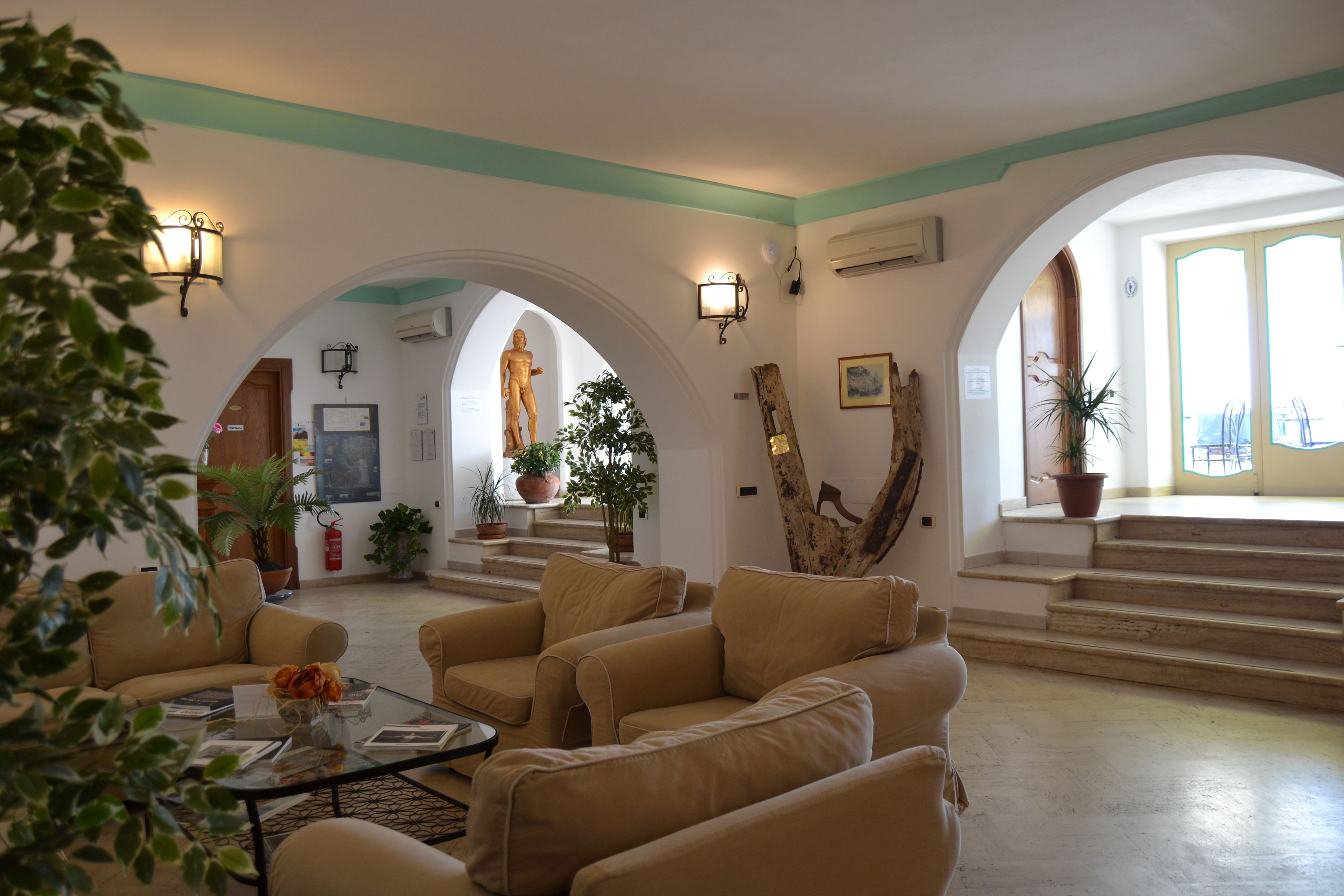 Zona saló àmplia amb sofàs beix, una taula de centre i plantes en testos. Hi ha portals arquejats que condueixen a altres habitacions, i una estàtua es veu al fons.