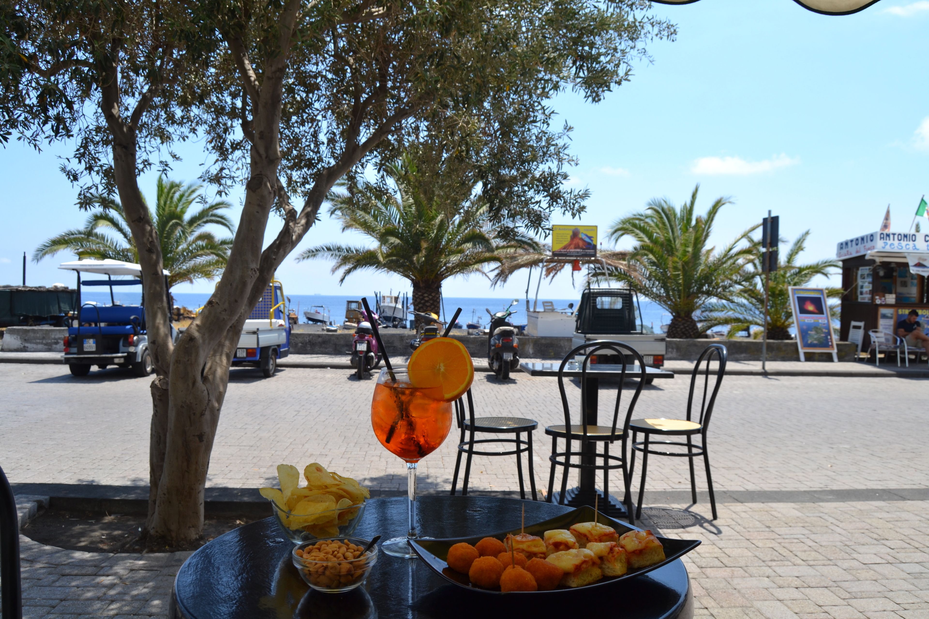 Una mesa con comida y bebidas, una palmera y vista al mar con vehículos al fondo.