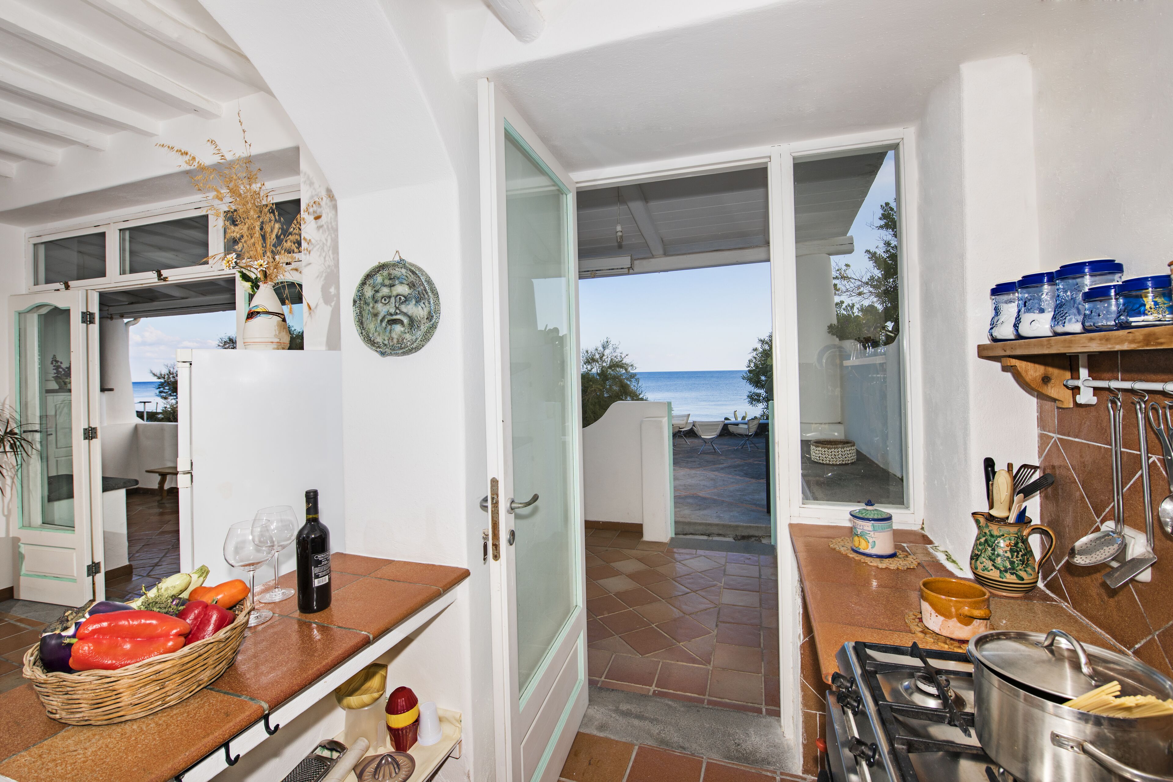 Una cocina con vista al mar, una cesta de mimbre llena de verduras, una botella de vino y utensilios de cocina.