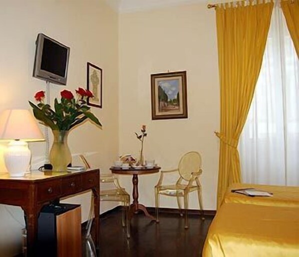 A hotel room with a bed, a desk with a lamp, a vase with flowers, a chair, and a television on the wall.