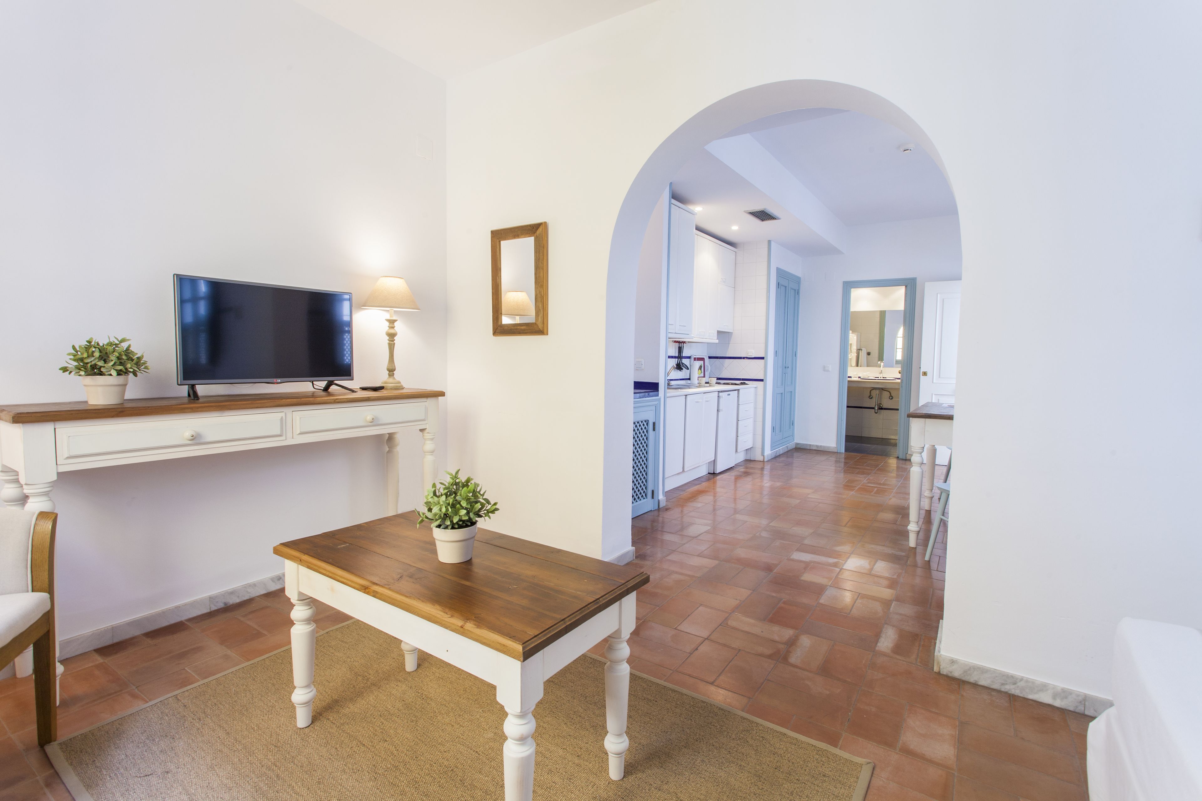 A modern living room with a TV, wooden table, and potted plants. An archway leads to a kitchen and dining area.