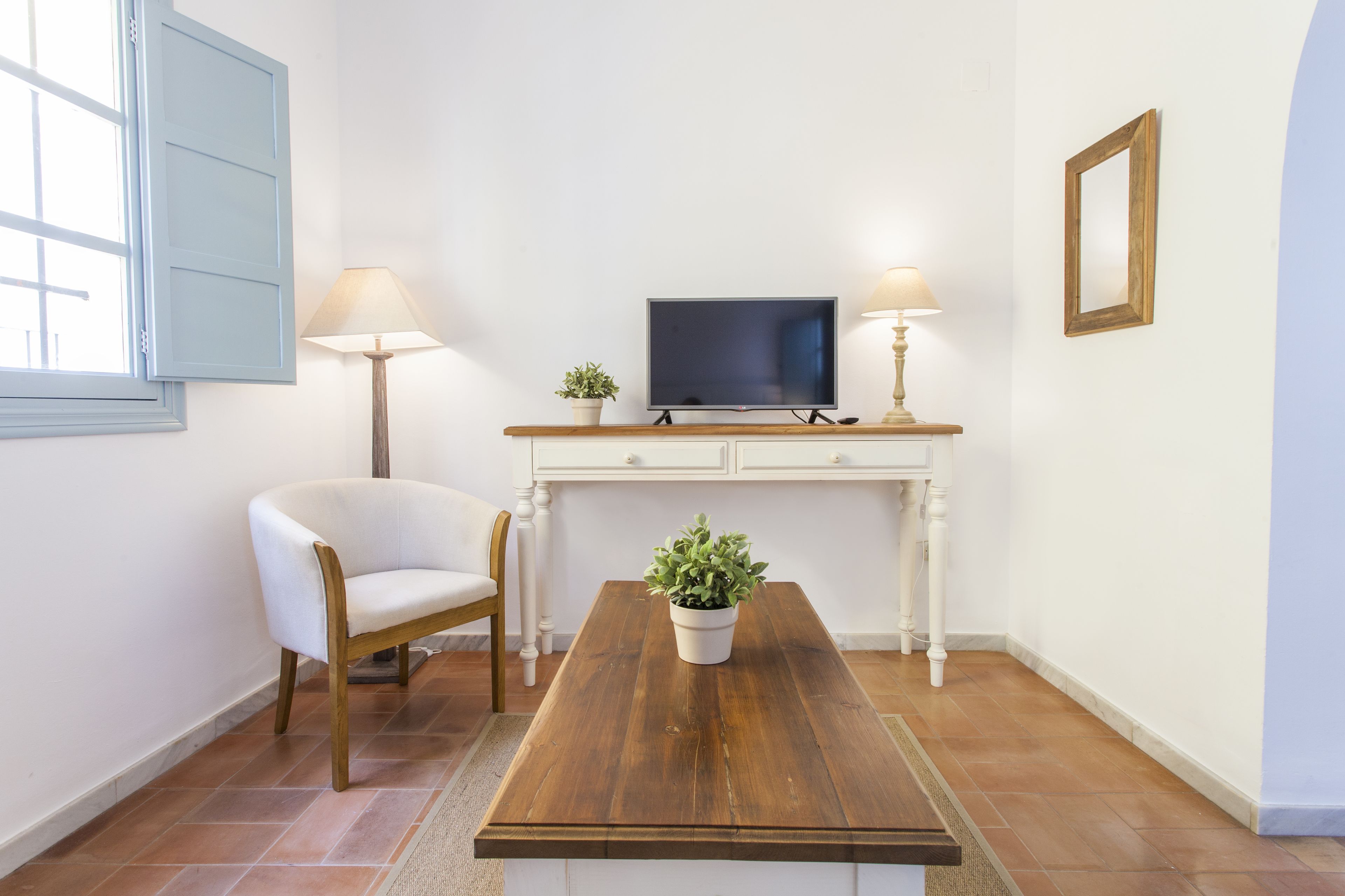 A room with a wooden coffee table, a white chair, a television on a white cabinet, and two lamps.