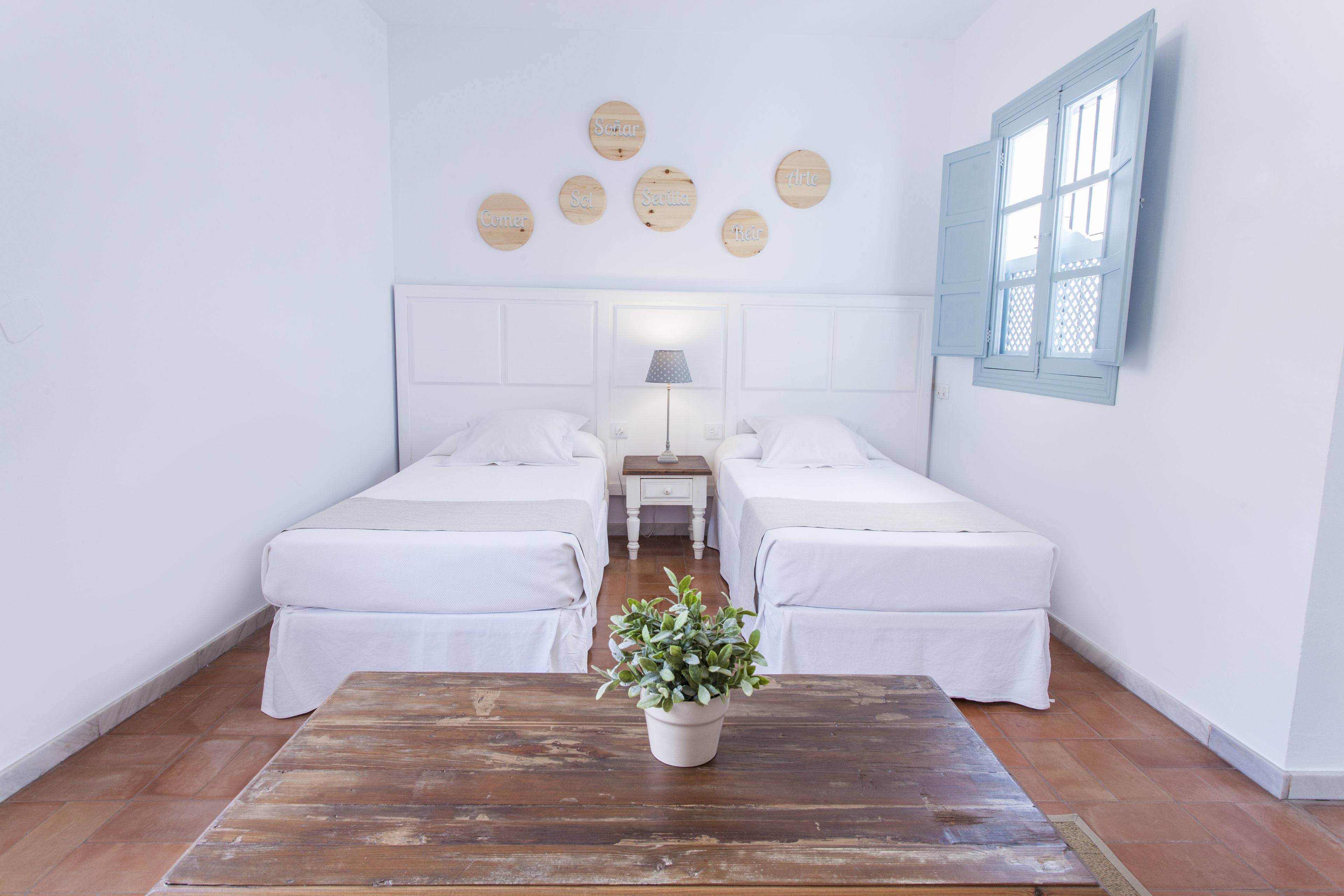 A room with two beds, a wooden table with a plant, a nightstand with a lamp, and a wall with circular decorations.
