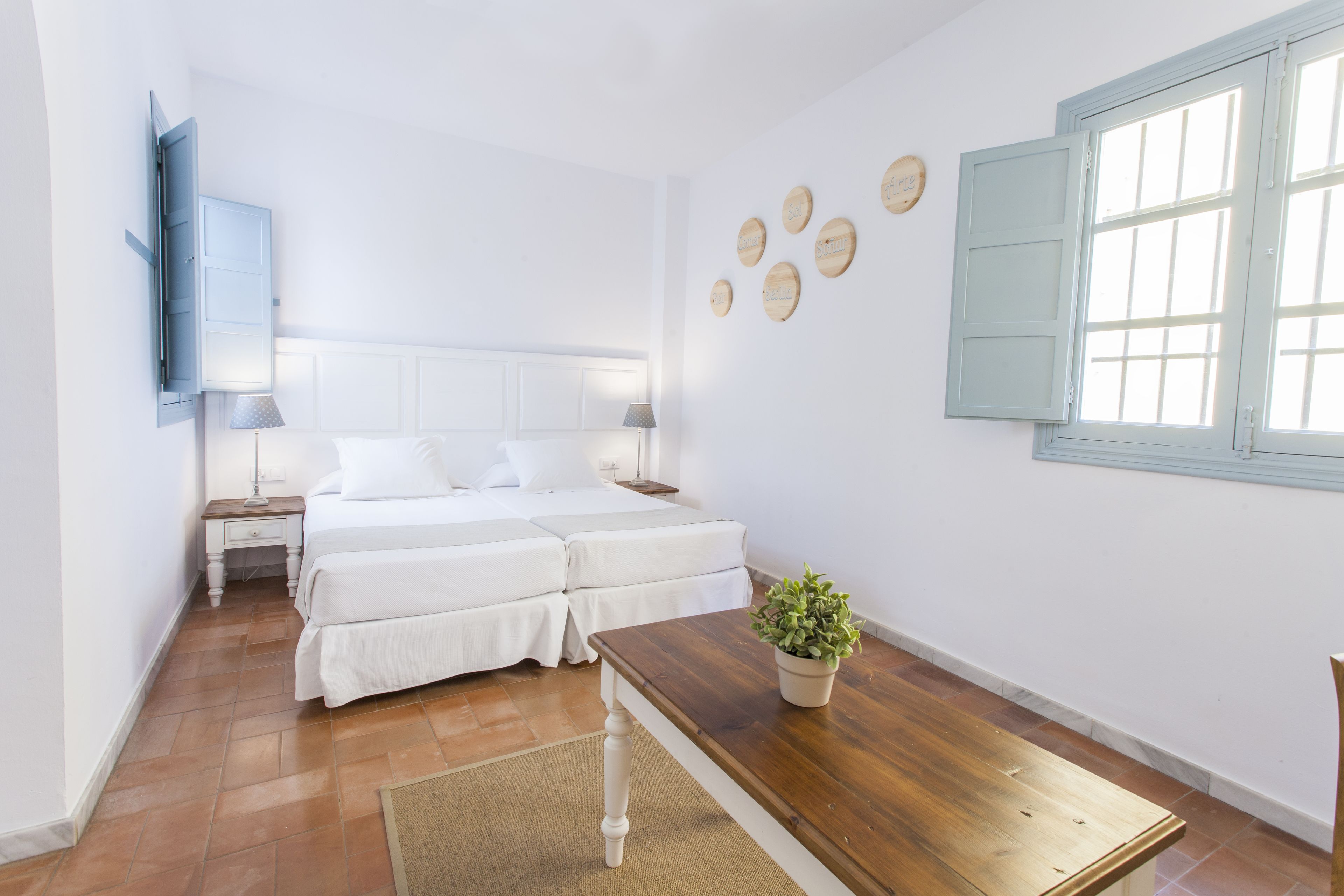 A bedroom with two beds, a wooden table, a potted plant, and a window with shutters.