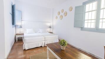A bedroom with two beds, a wooden table, a potted plant, and a window with shutters.