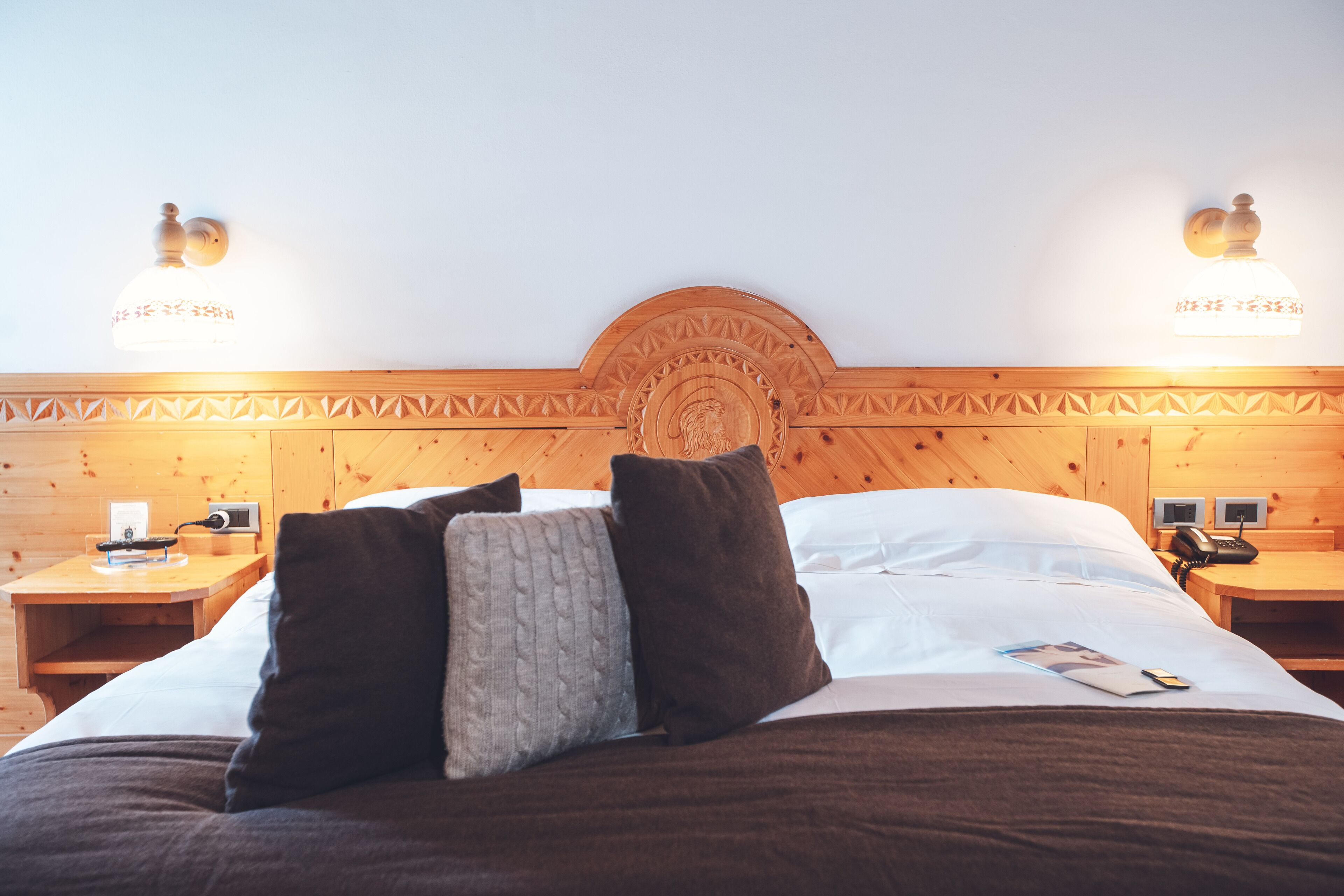 Una cama bien hecha con cabecera de madera, dos mesitas de noche con lámparas y un teléfono instalado en la pared.