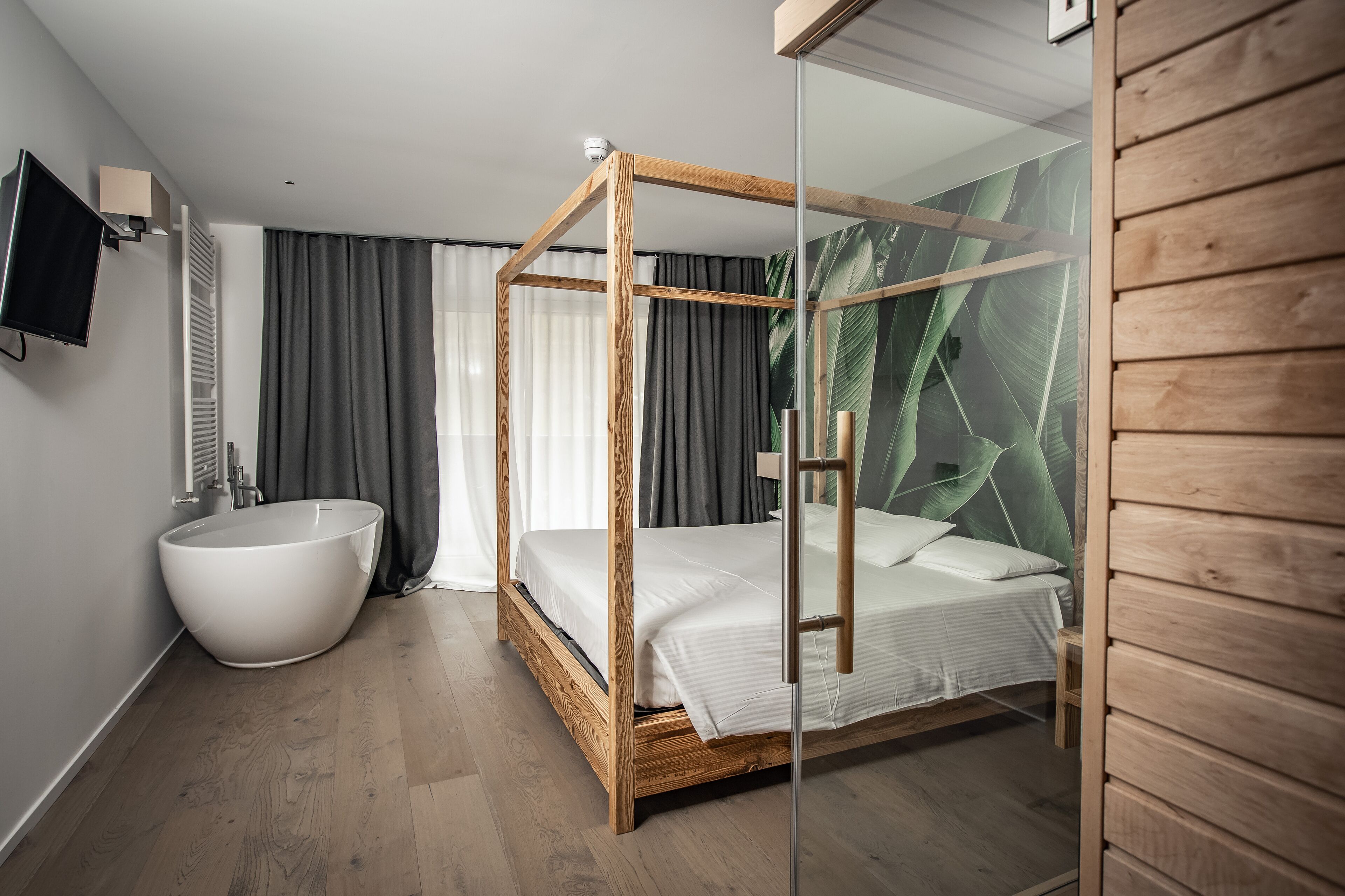A modern bedroom with a wooden four-poster bed, a white bathtub, and a glass partition with a tropical leaf design.