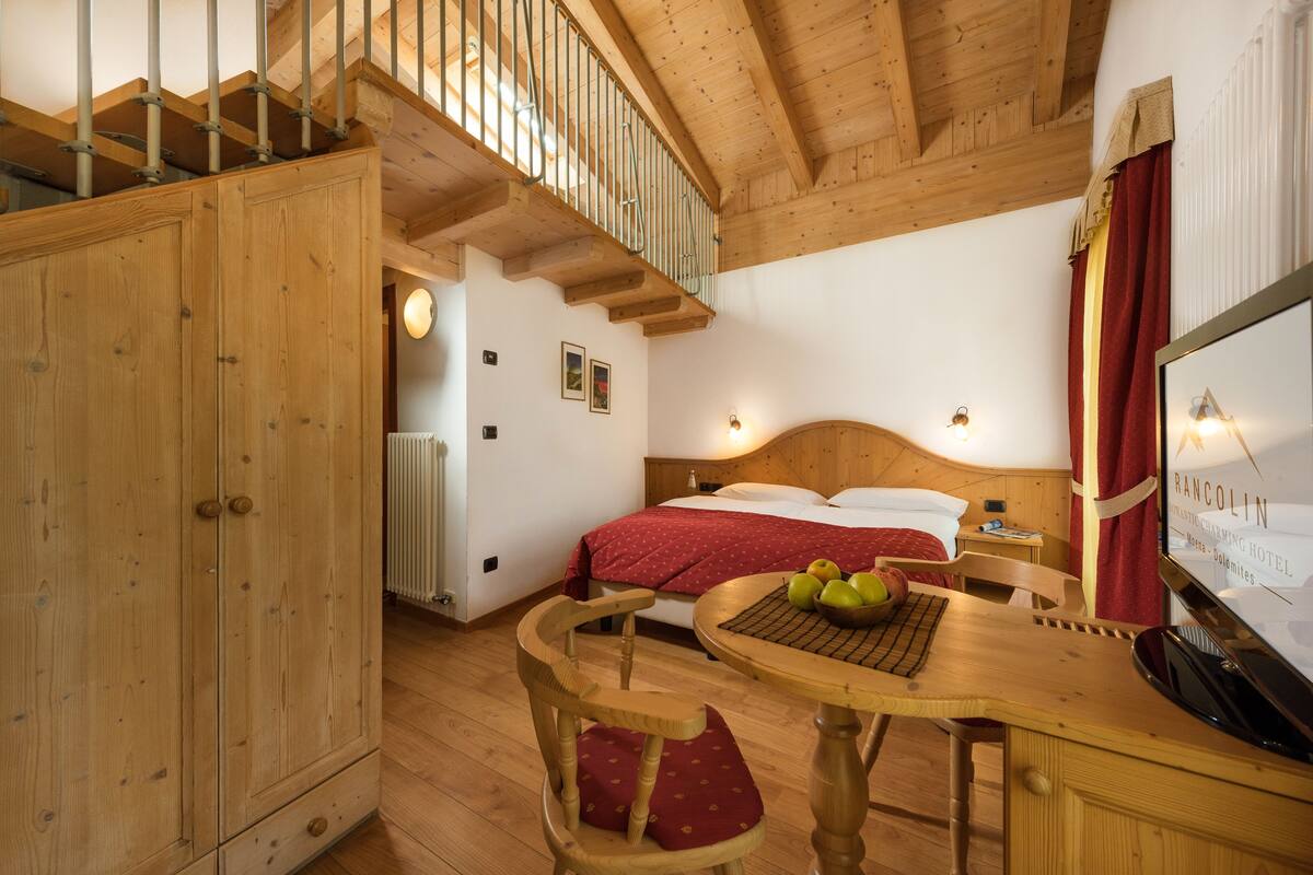 Ein Holzinnenraum mit einem Bett, einem Schreibtisch mit Äpfeln, einem Sessel und einem Fernseher, der Hotelinformationen zeigt.
