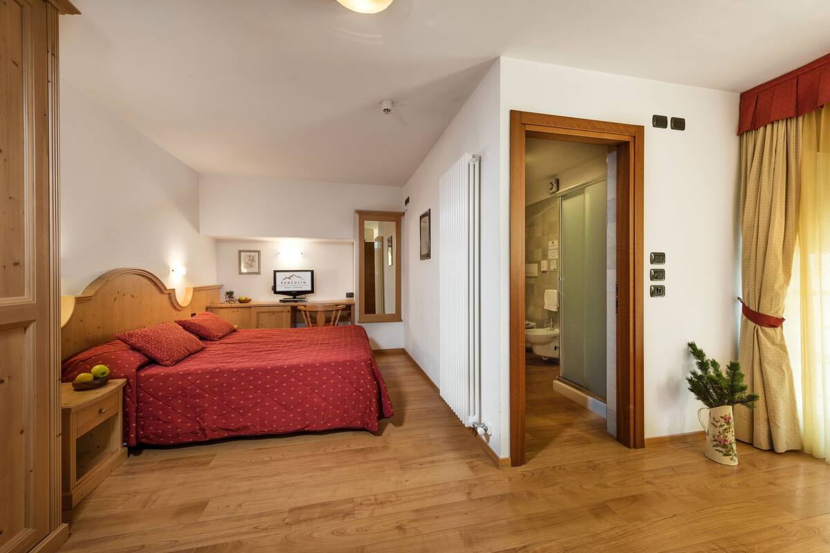 Ein Schlafzimmer mit einem hölzernen Bett, einem Nachttisch mit Obst, einem Schreibtisch mit einem Computer und einem durchgehenden Türspalt sichtbaren Badezimmer.