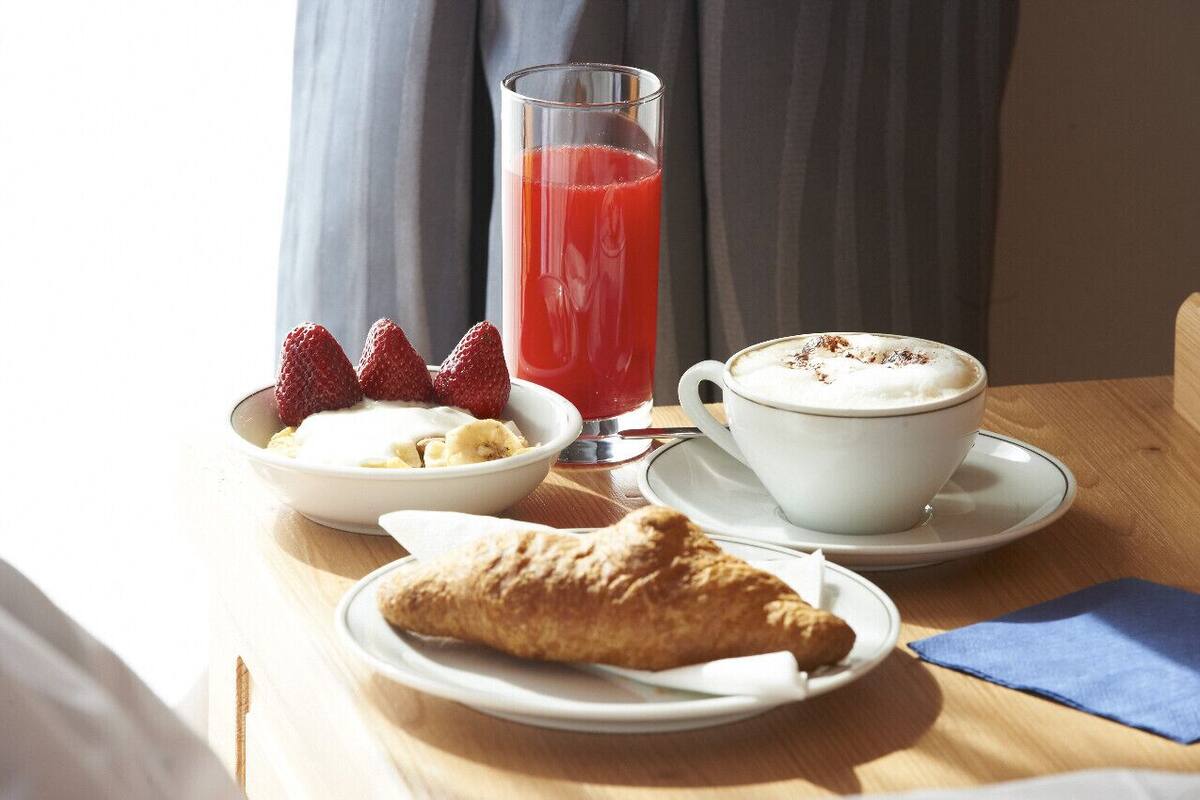 A breakfast setting with a croissant, a cup of coffee with foam, a bowl of yogurt with strawberries, and a glass of red juice on a wooden table.