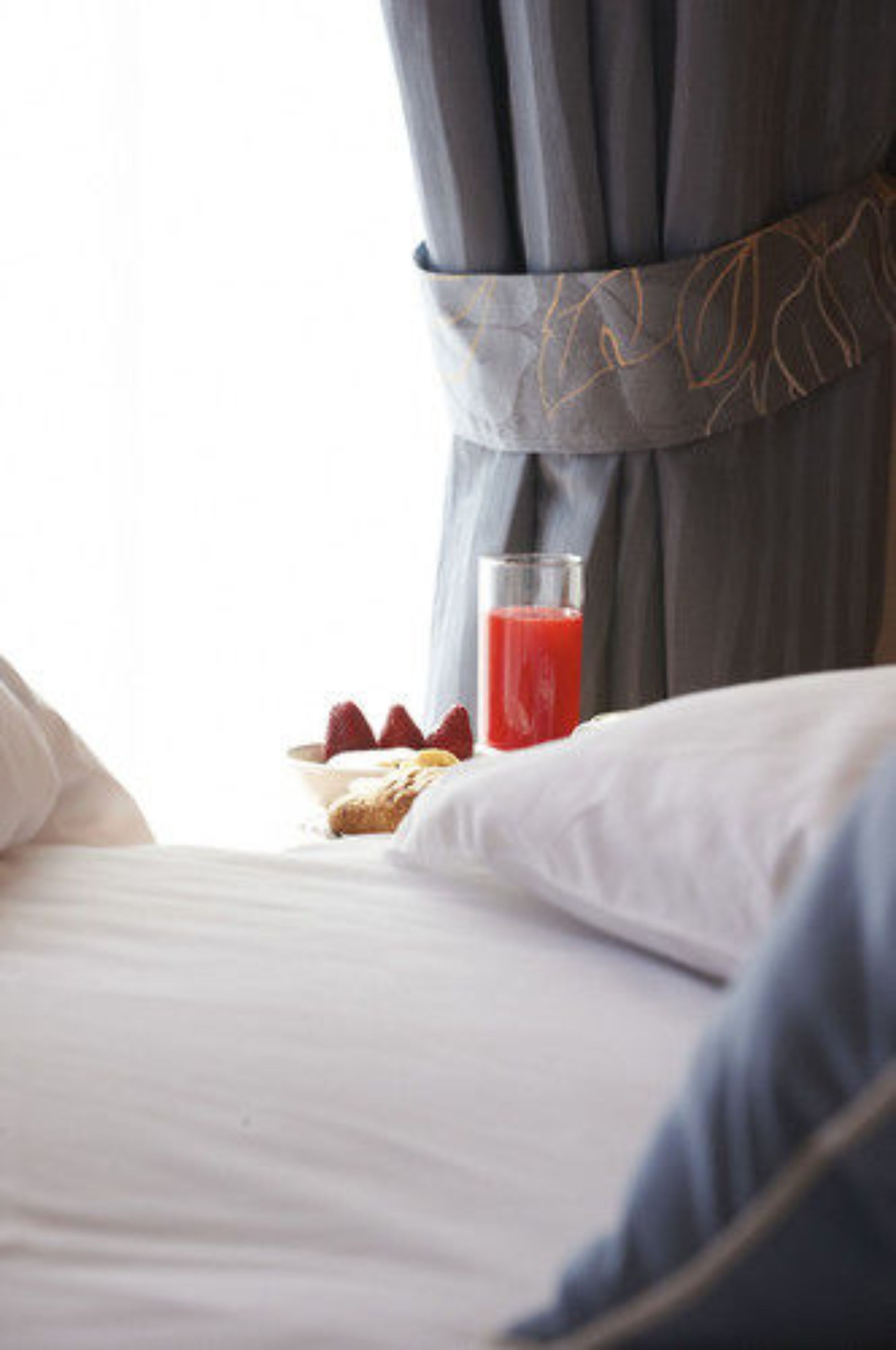Una cama bien hecha con ropa blanca, un vaso de jugo de frutos rojos y un cuenco con fresas al lado.