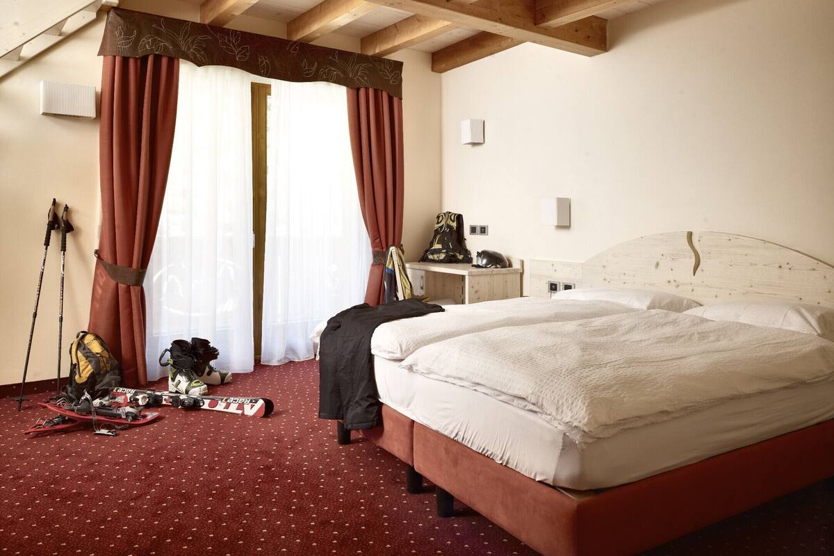 A hotel room with a wooden ceiling, a bed with white bedding, a desk with a chair, and ski equipment on the floor.