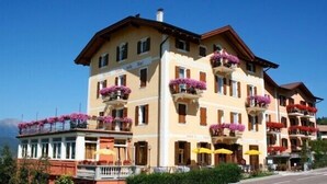 A multi-story hotel with balconies adorned with flowers, outdoor seating, and a mountain backdrop. - Stella delle Alpi (Ronzone)