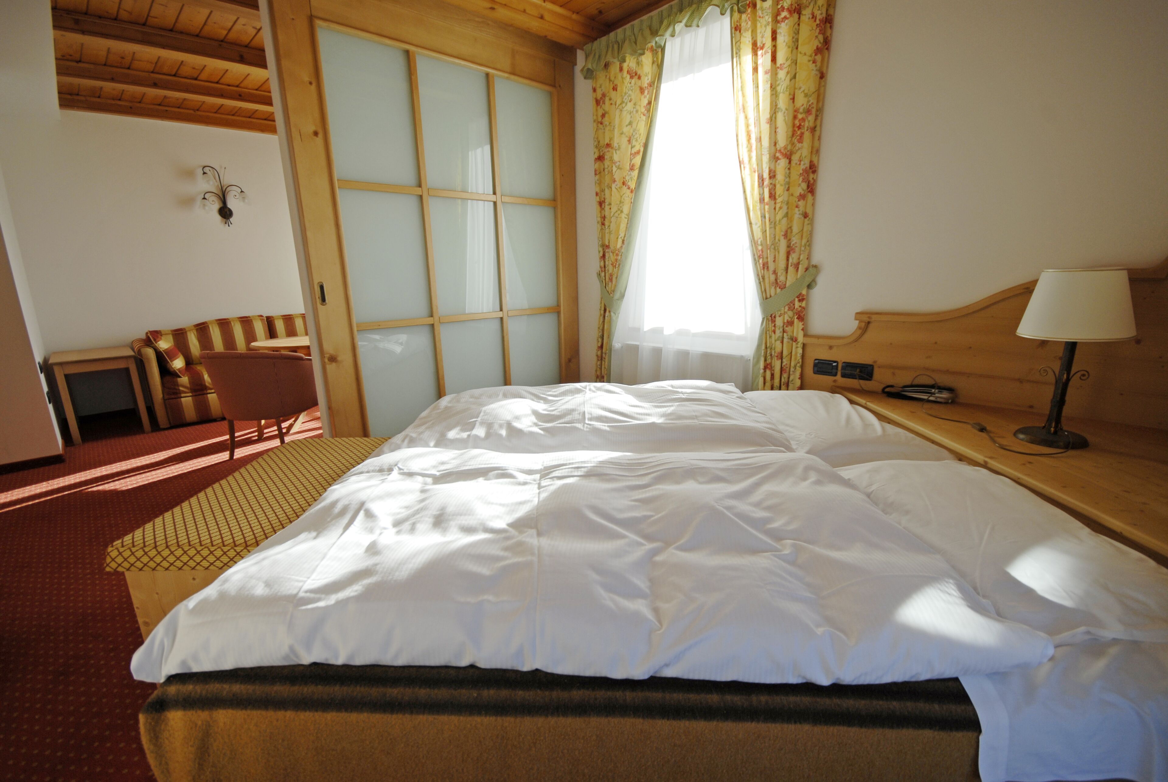 Camera d'albergo con un letto grande, una testiera in legno, un comodino con una lampada, una piccola scrivania con una sedia e una finestra con tende.