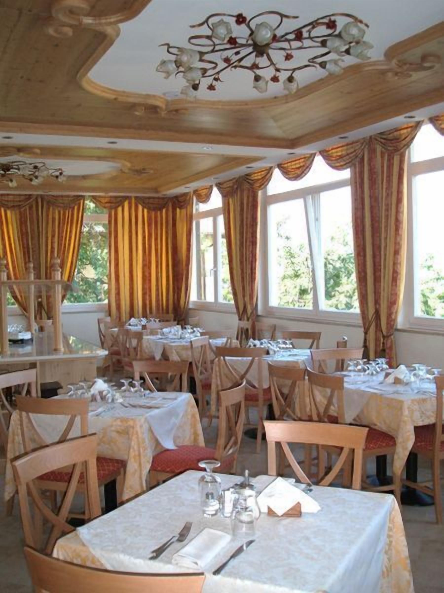 Un restaurant doté de nombreuses tables et chaises, de grandes fenêtres avec des rideaux et d’un lustre.