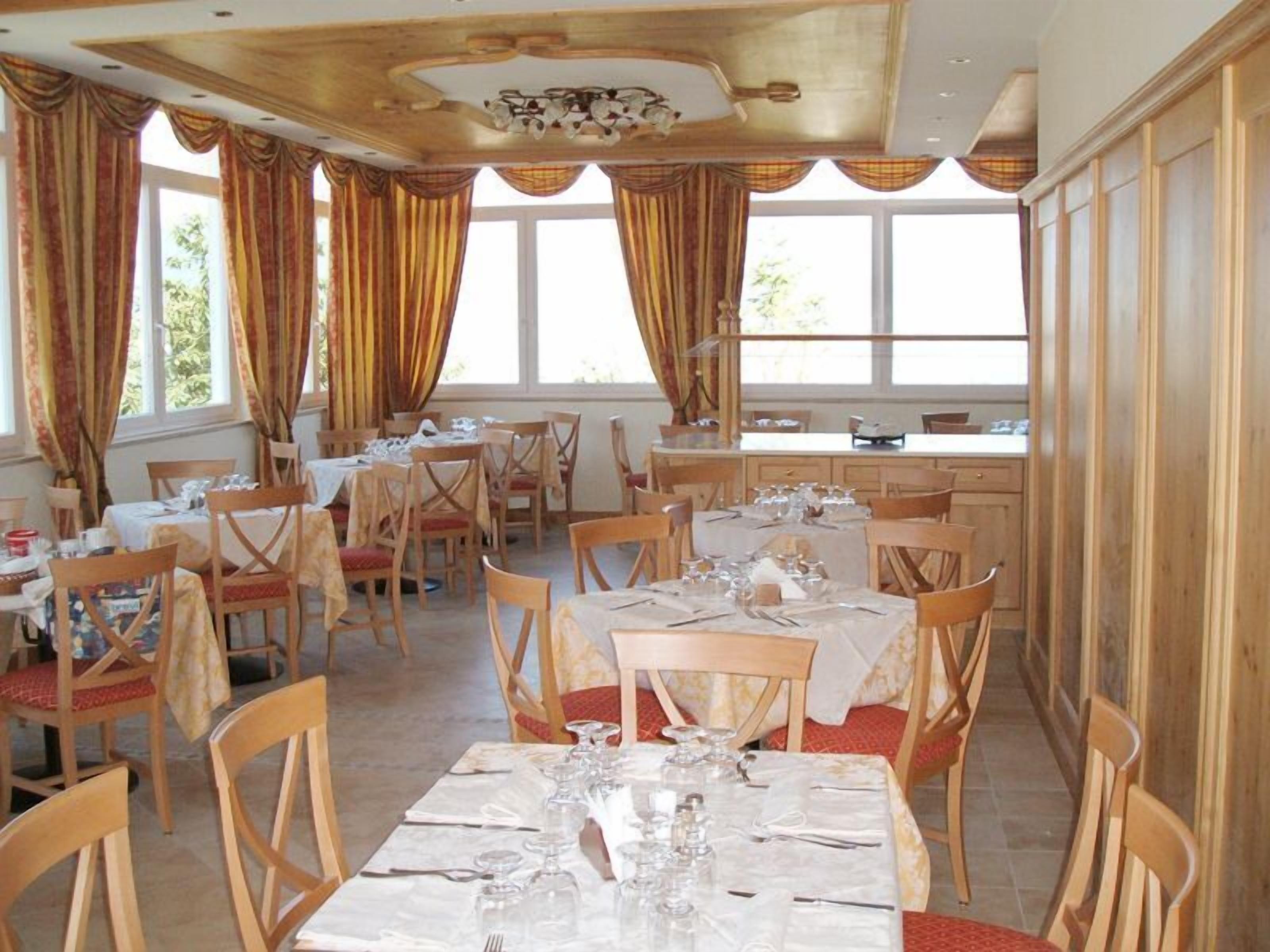 Un comedor con varias mesas dispuestas para el servicio, sillas de madera y amplias ventanas con cortinas.