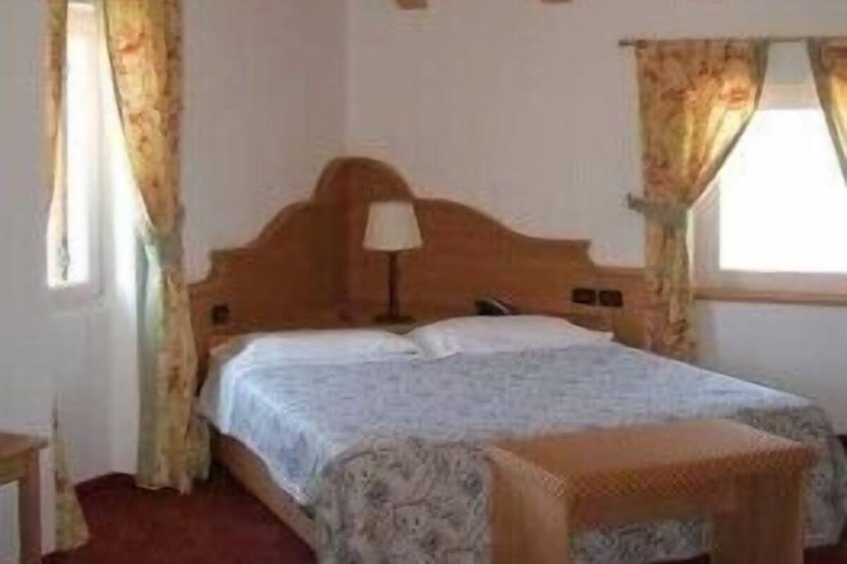 Un dormitorio con techo de madera, dos ventanas con cortinas, una cama con colcha estampada y una mesita de noche.
