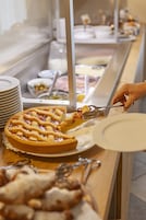 Mostrador de buffet con diversos alimentos y una mano usando pinzas para servir una porción de tarta.