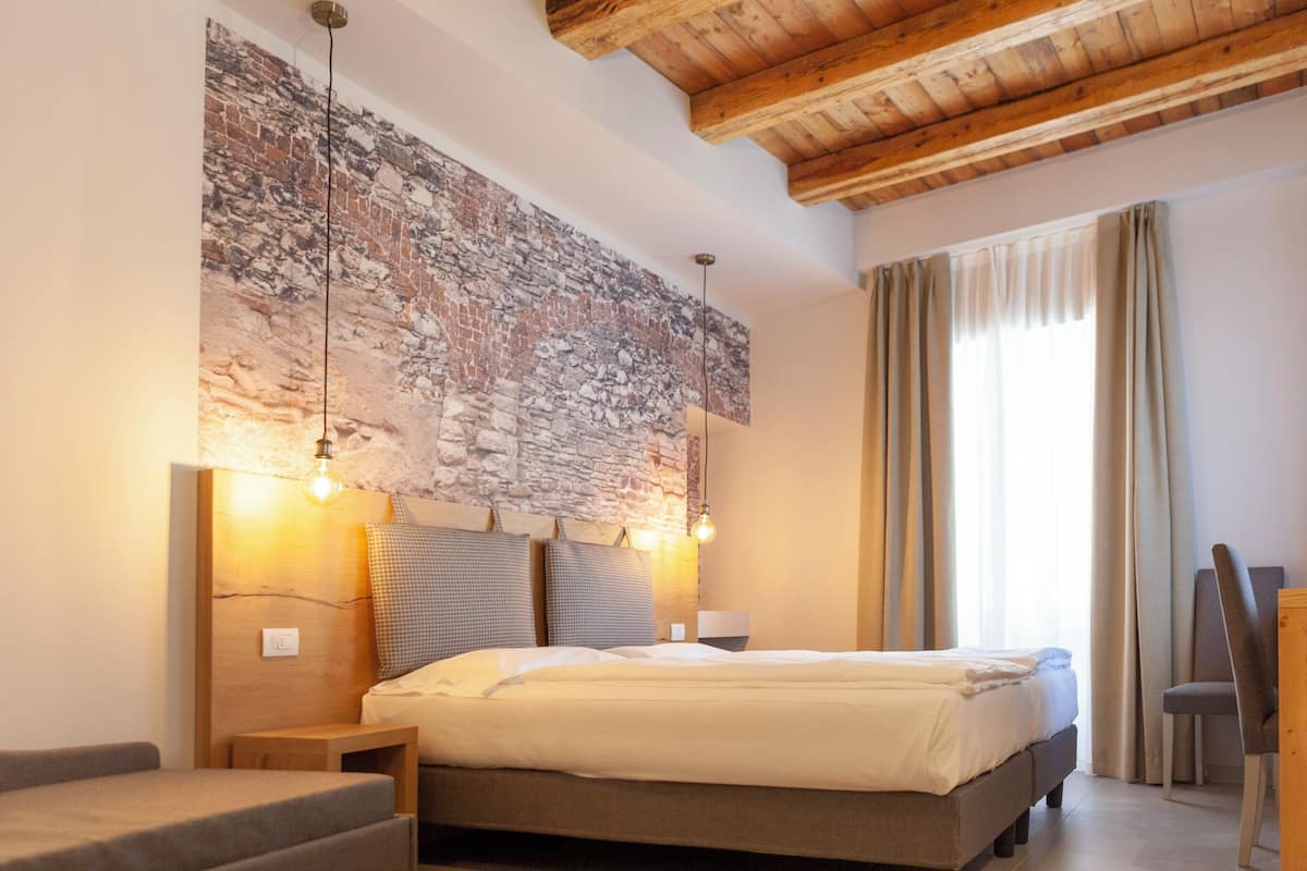 Un dormitorio con pared de piedra, techo de madera, cama con cabecera, lámparas de noche, una silla y ventana con cortinas.