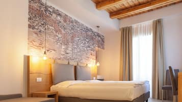 Un dormitorio con pared de piedra, techo de madera, cama con cabecera, lámparas de noche, una silla y ventana con cortinas.