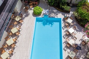 Una piscina en la azotea con superficie de agua azul cristalina, rodeada de zonas de estar al aire libre con sillas y mesas de mimbre, y un pequeño arbusto verde.