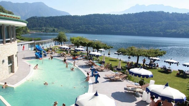 Ein Poolbereich mit Wasserrutsche, Liegestühlen, Sonnenschirmen und Blick auf einen See und Berge.