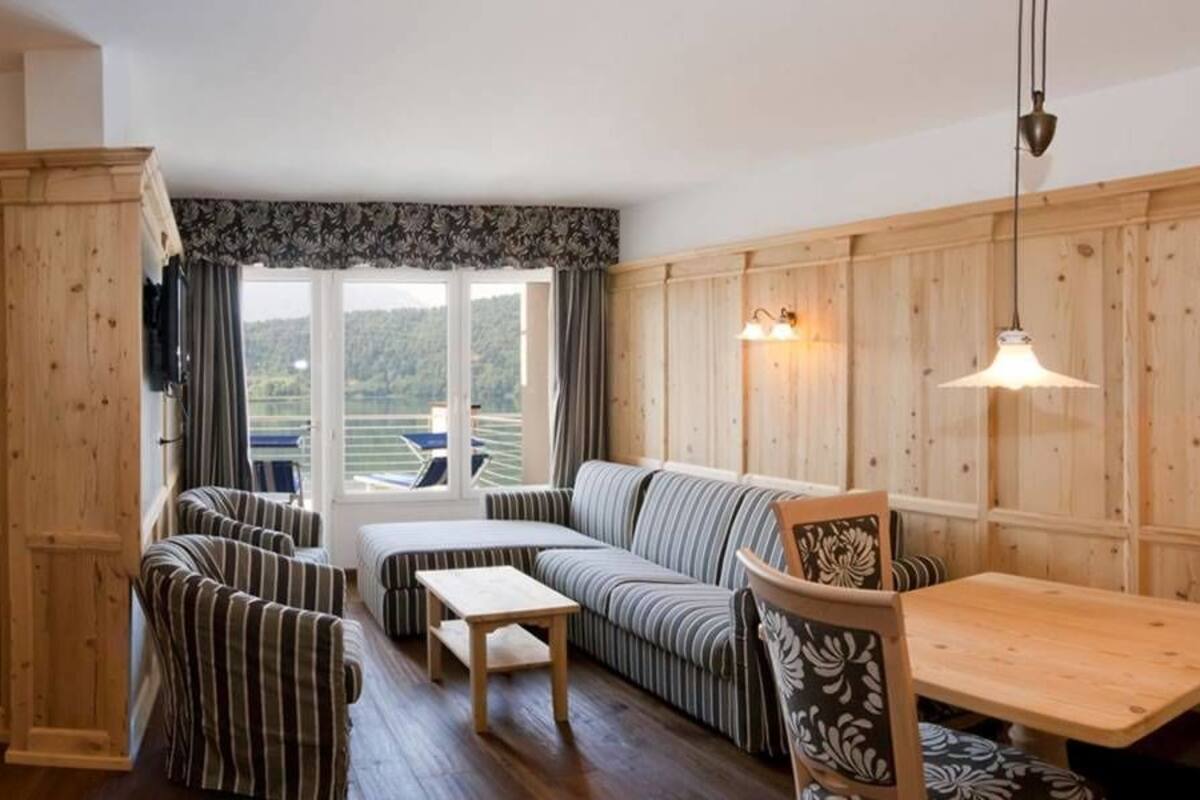 A cozy room with a wooden interior, a sofa set, a small table, and a view of a green landscape through large windows.