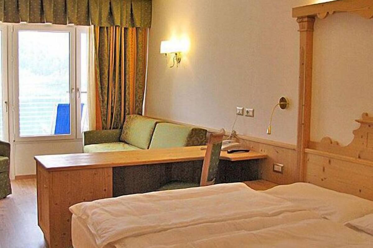 A hotel room with a bed, a desk, a chair, a window with curtains, and a wall-mounted lamp.