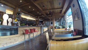 A restaurant interior with wooden ceiling beams, a bar area with bottles and glasses, red bar stools, and wooden tables and benches. - Hotel Arnica (Pinzolo)