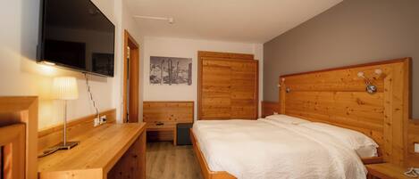 A bedroom with a wooden bed, a TV mounted on the wall, a wooden desk, and a wooden wardrobe.