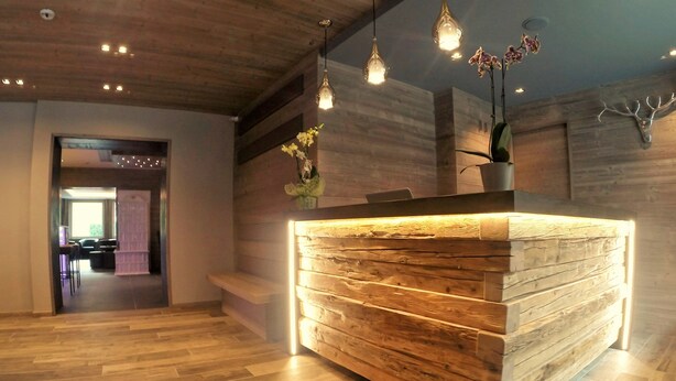A reception desk with a wooden finish and integrated lighting, a wooden ceiling, and hanging pendant lights. - Hotel Arnica (Pinzolo)