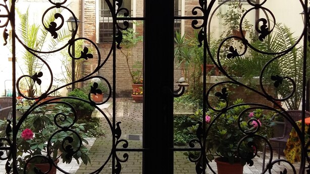 Una reja de hierro forjado con diseños intrincados, que da acceso a un patio con macetas y un camino de ladrillo.