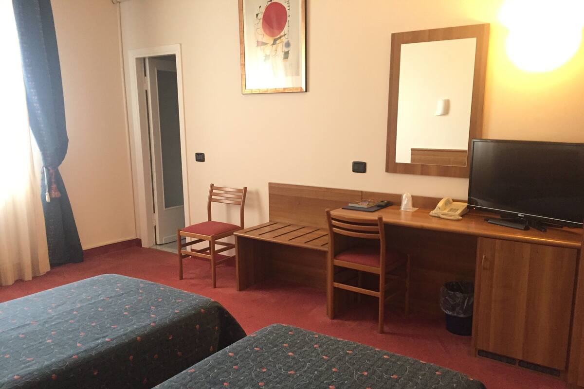 A hotel room with two single beds, a wooden desk with a chair, a television, and a small table.