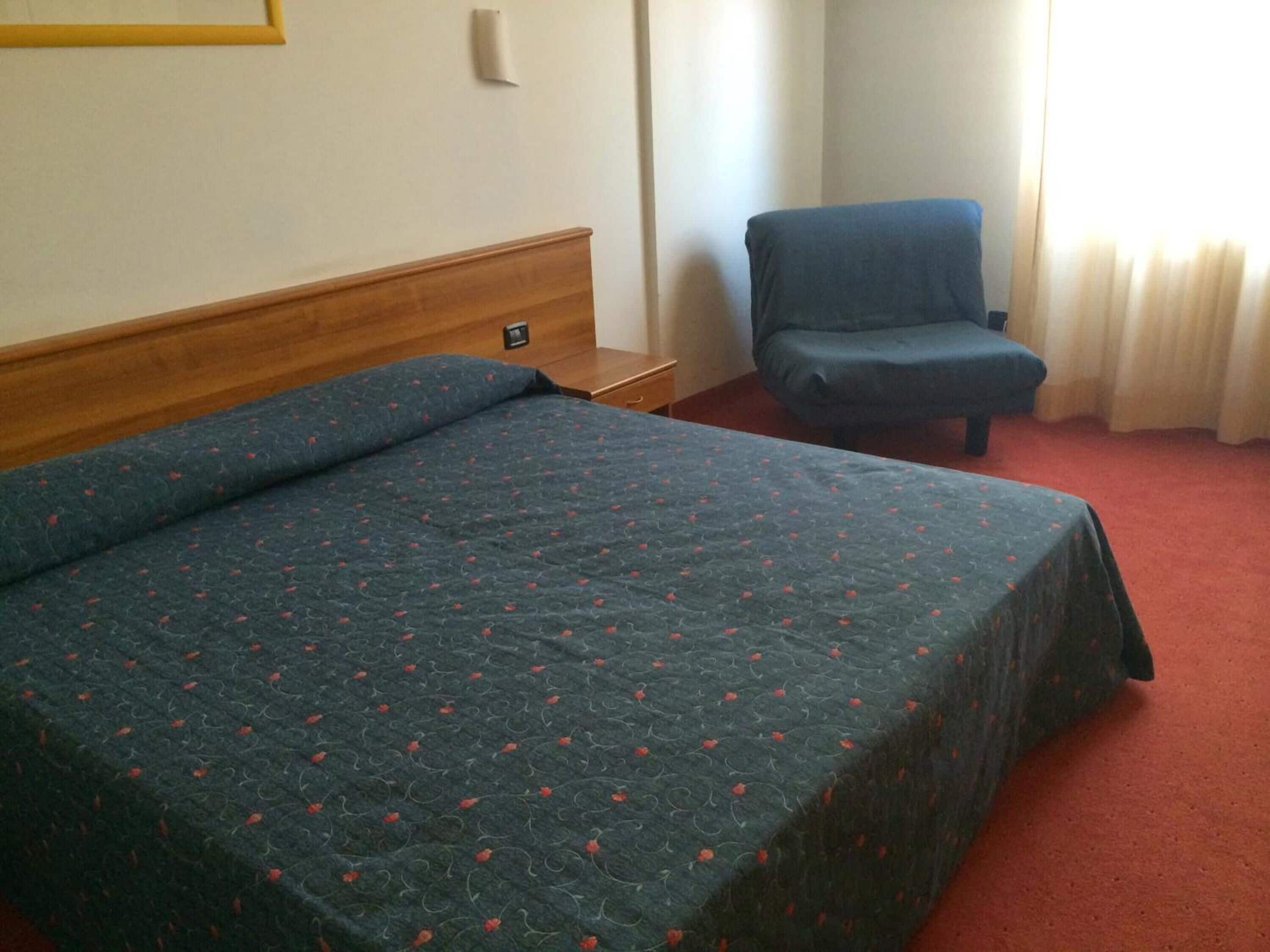 Une chambre d’hôtel avec un lit, une chaise, une tête de lit en bois, une lampe fixée au mur et une fenêtre avec des rideaux.