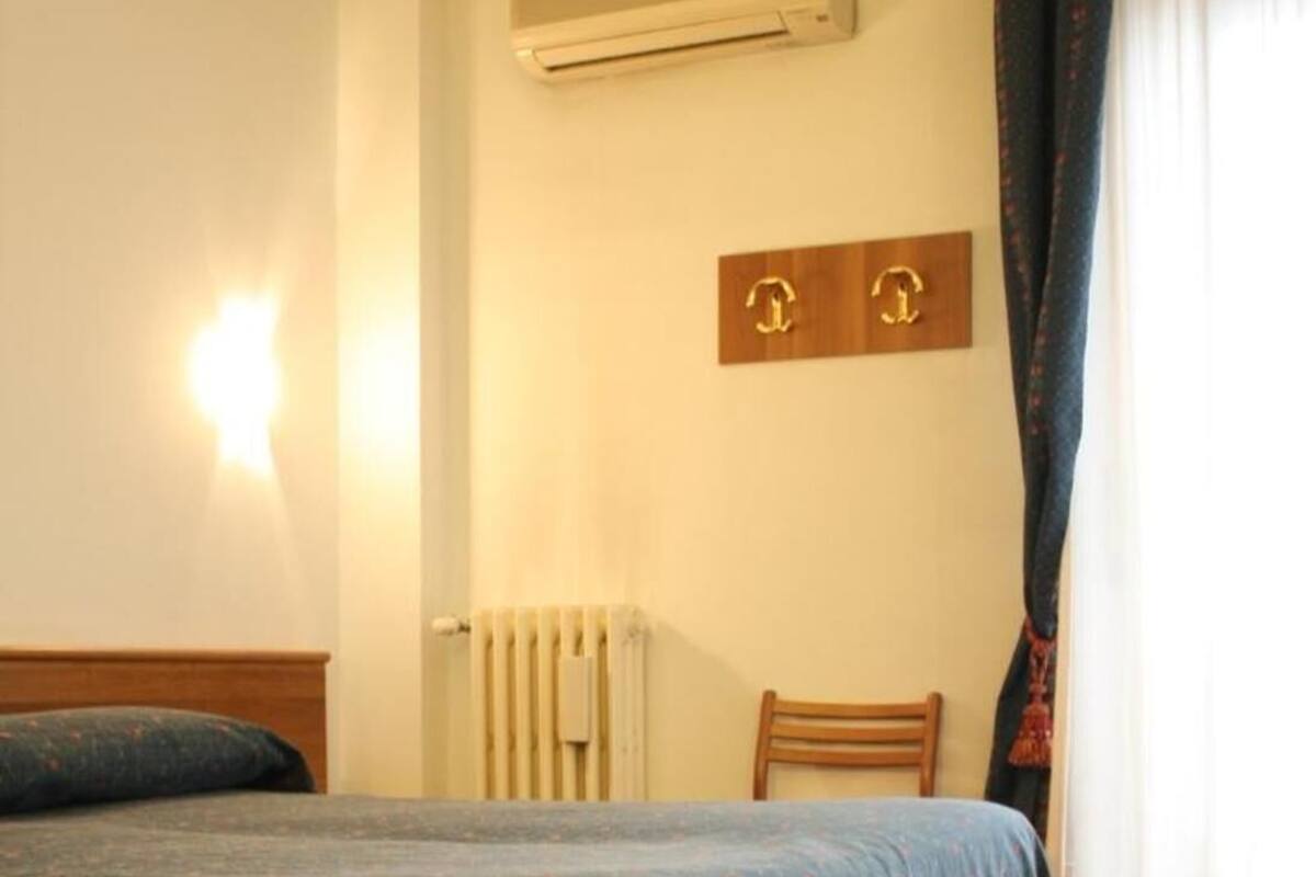 A hotel room with a bed, a radiator, an air conditioning unit, a wooden headboard, a chair, and a window with curtains.